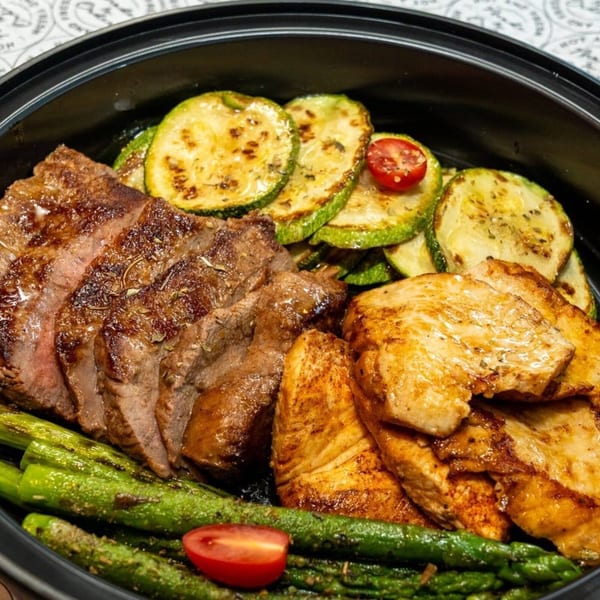 Grilled steak, chicken, zucchini, and asparagus with cherry tomatoes in a black bowl.