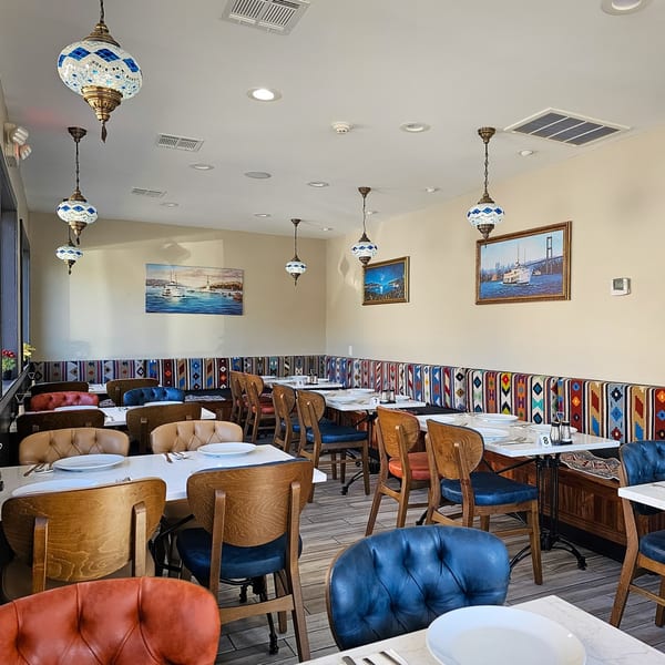 A brightly lit dining room with patterned banquettes, wooden tables, and colorful chairs under ornate hanging lamps.