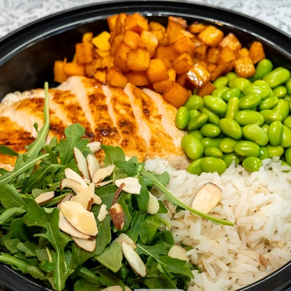 Grilled chicken, rice, edamame, sweet potatoes, and arugula with almonds in a black bowl.