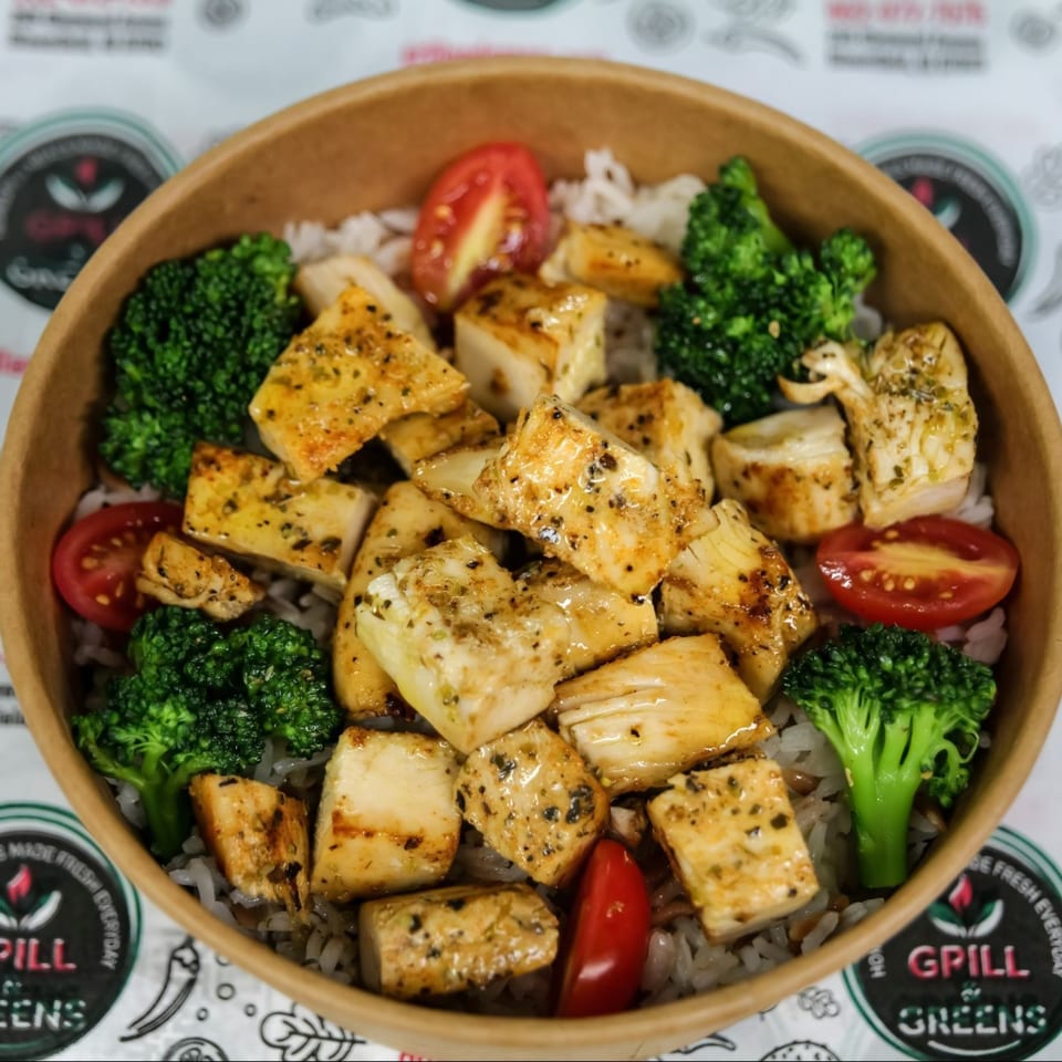 Grilled chicken, broccoli, and cherry tomatoes served over rice in a brown paper bowl.