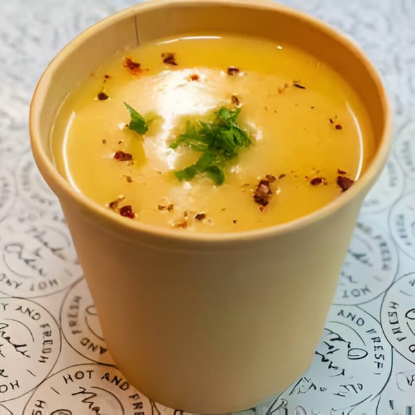 Creamy yellow soup in a brown paper cup, garnished with green herbs and red pepper flakes.