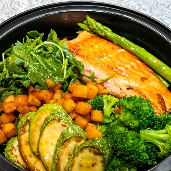 Grilled salmon with asparagus, broccoli, zucchini, butternut squash, and arugula in a black bowl.